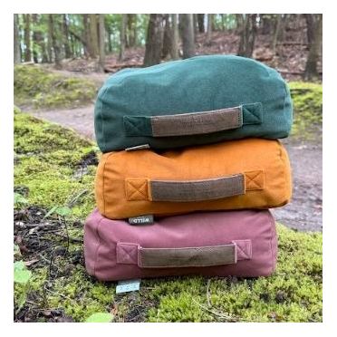 Trzy poduszki do medytacji ułożone jedna na drugiej – zielona, musztardowa i różowa – stoją na pokrytym mchem pniu w lesie, w tle drzewa i ścieżka.